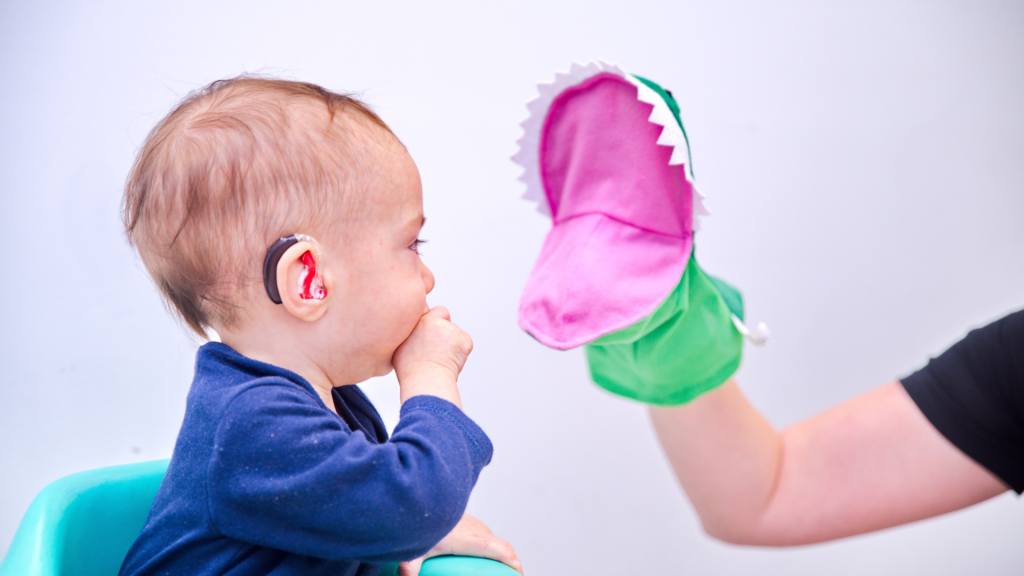 Baby with hearing aid looking at hand puppet in speech therapy session.