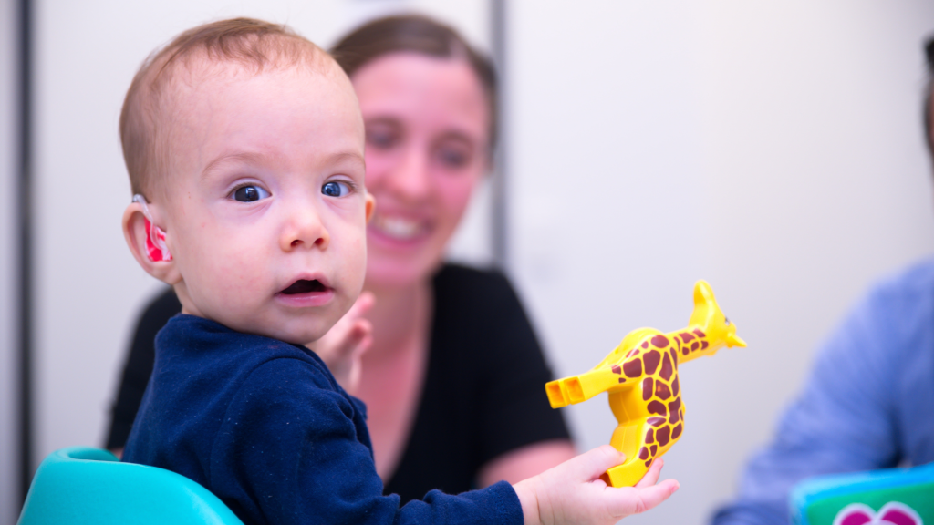 Speech pathologist and child with hearing aid holding a toy giraffe looking at camera.