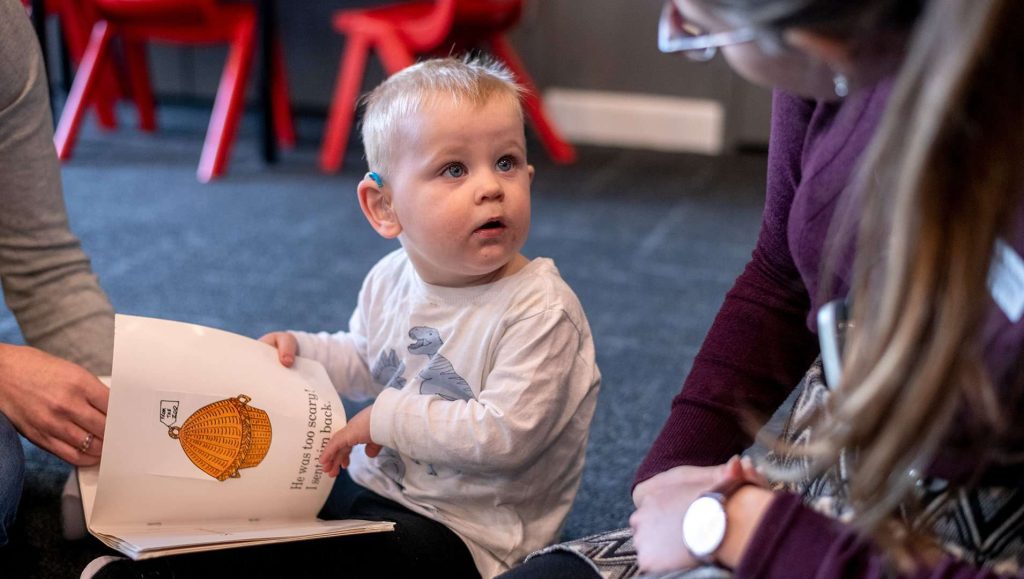 Toddler with hearing aid reading picture book with speech pathologist.