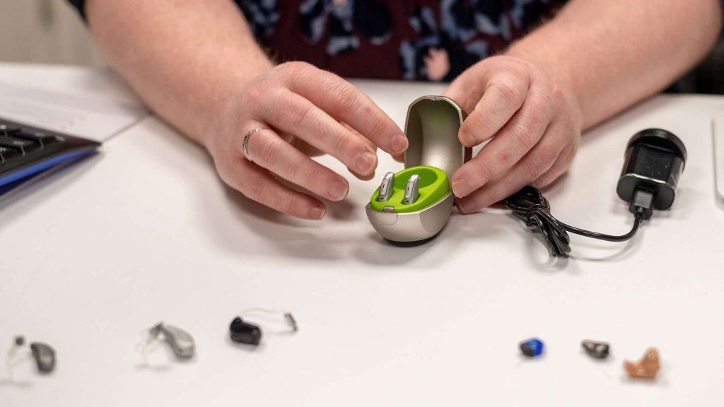 A person’s hands holding a pair of hearing aids in a case, with several other hearing aid options in front of them.